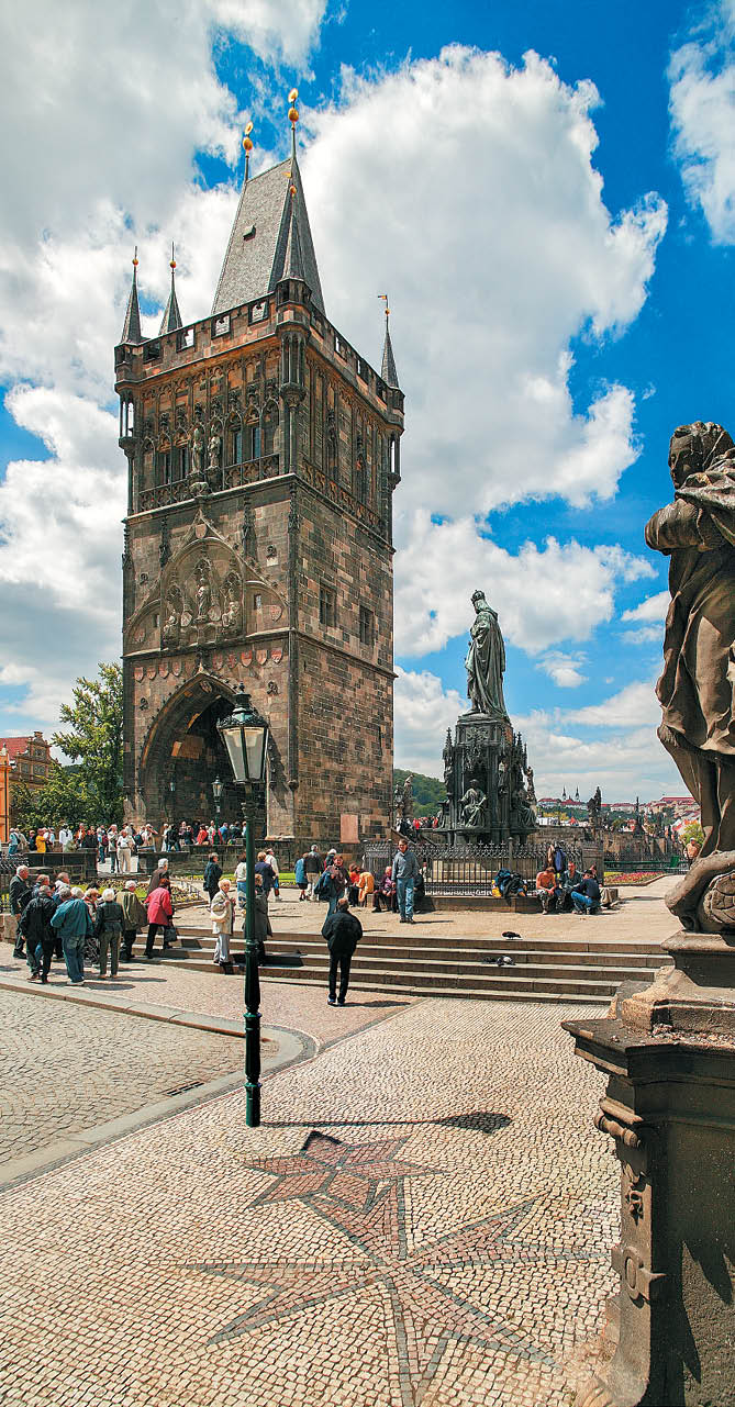 Historick v   s gotickou architekturou stoj  na n m st  s dla bou a je obklopena lidmi za slune n ho dne s modrou oblohou a mraky.  Popis generovan  AI