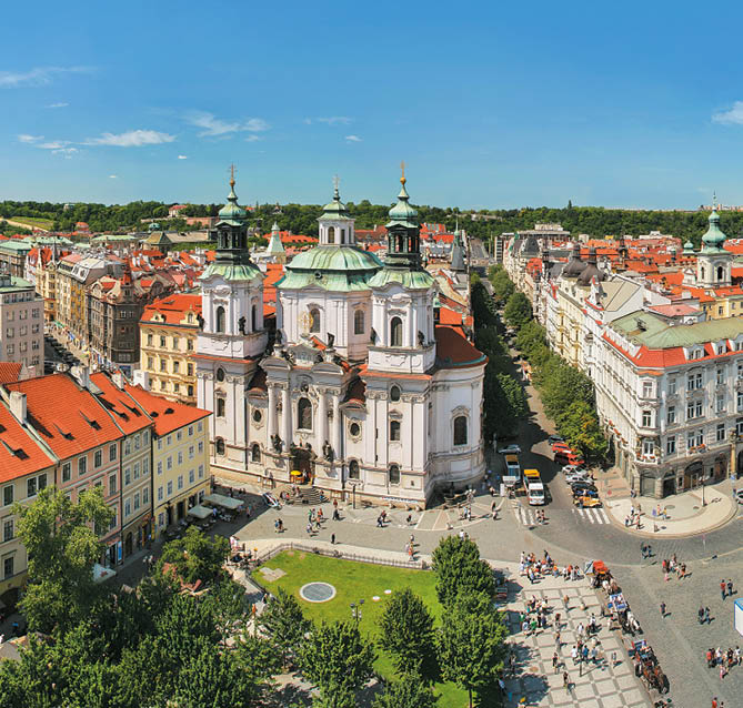 Barokn kostel s v  emi obklopen  historick mi budovami a lidmi na n m st  za slune n ho dne.  Popis generovan  AI