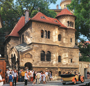 Historick budova s  ervenou st echou stoj  u ru n  ulice, kde se shroma  uje skupina lid  a proj  d  staro itn  auto.  Popis generovan  AI