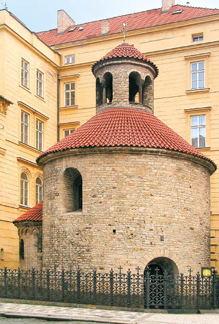 Kulat kamenn  stavba s  ervenou st echou a malou v  i kou stoj  v uzav en m prostoru mezi  lut mi budovami.  Popis generovan  AI