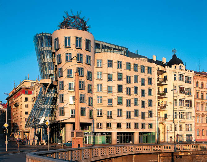 Budova s neobvyklou architekturou zahrnuj c zkosen  a zak iven  tvary stoj  u  eky za jasn ho dne.  Popis generovan  AI