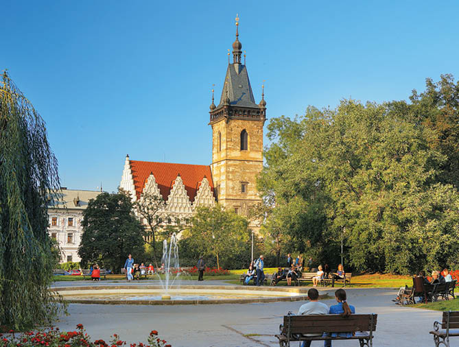 V parku s font nou sed lid  na lavi k ch a v pozad  je historick  budova s v    a  ervenou st echou za jasn ho modr ho nebe.  Popis generovan  AI