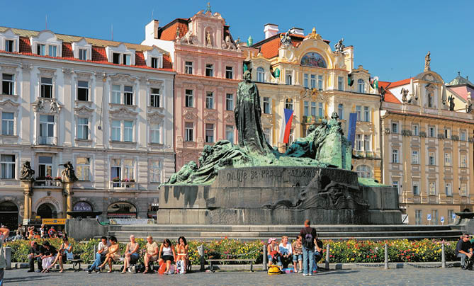 V historick m n m st s barevn mi budovami stoj  velk  bronzov  socha obklopen  lidmi sed c mi na lavi k ch a proch zej c mi se.  Popis generovan  AI