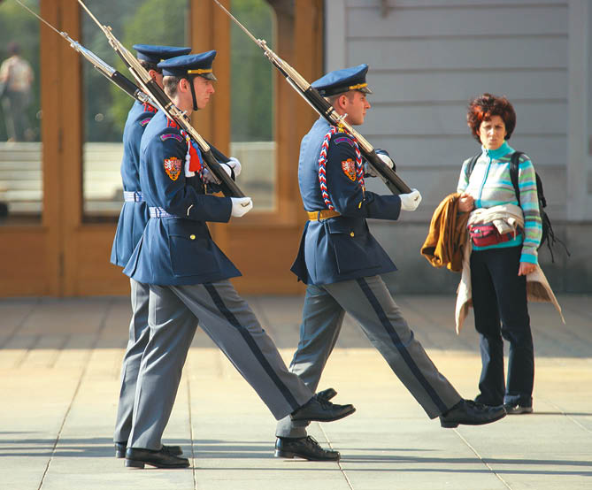 T i lid v uniform ch s pu kami vykon vaj  slavnostn  pochod na dla b  p ed budovou a dal   osoba stoj  vedle nich s batohem a bundou v ruce.  Popis generovan  AI