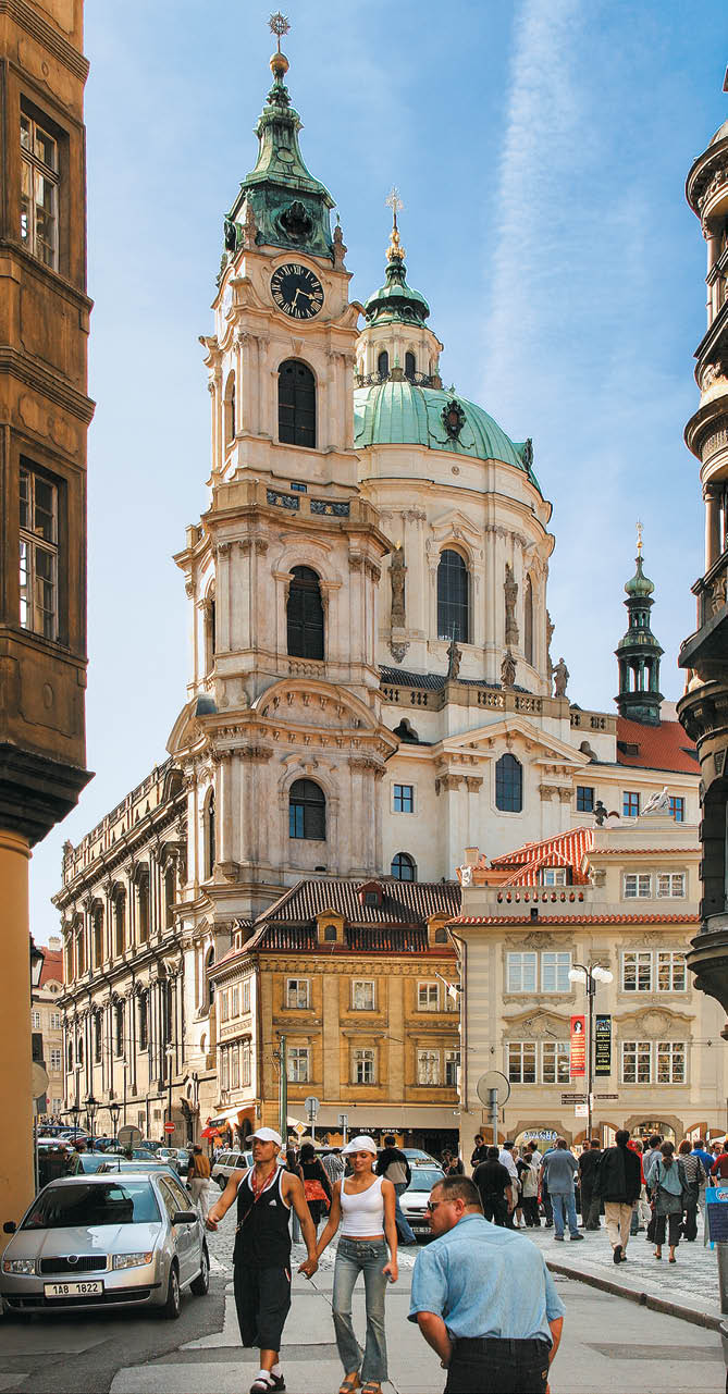 V historick m m stsk m prost ed s barokn  architekturou stoj  lid  na ulici vedle zaparkovan ch aut a velk  kosteln  v  e s hodinami a zelenou kopul .  Popis generovan  AI
