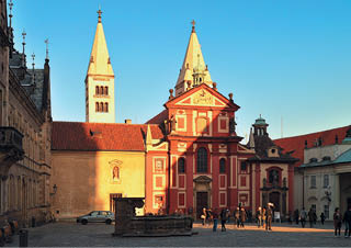 N m st s historickou architekturou v etn  dvou v    a  erven  budovy osv cen  sluncem, kde se pohybuje n kolik lid .  Popis generovan  AI