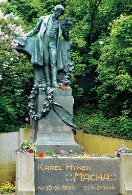 Socha osoby v historick m od vu dr c  kv tiny stoj c  na podstavci s n pisem Karel Hynek M cha obklopen  zelen  a kv tinami.  Popis generovan  AI