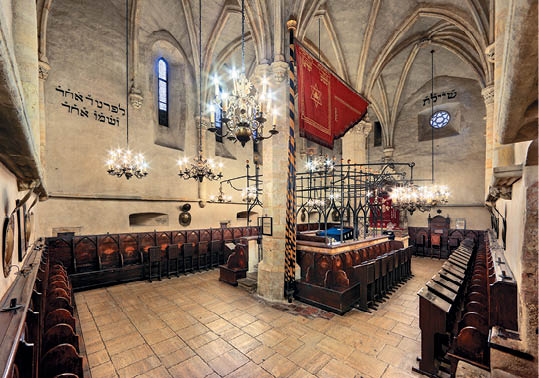 Interi r historick synagogy s d ev n mi lavicemi, kamennou podlahou, gotick mi klenbami a z v sn mi lustry.  Popis generovan  AI