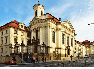 Barokn kostel s b l mi zdmi a  ervenou st echou stoj  u ru n  ulice, kde n kolik lid  p ech z  p es p echod a zaparkovan  jsou tam auta.  Popis generovan  AI