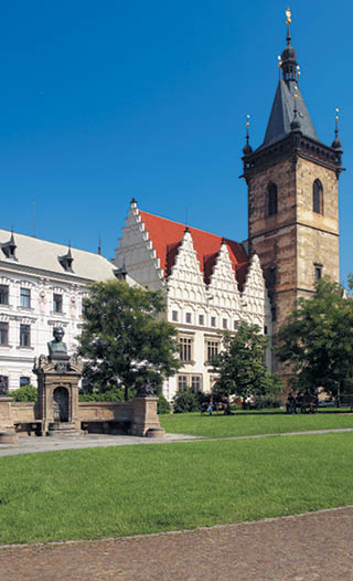 Historick budova s v    a sochou na n m st  s tr vn kem a stromy za slune n ho dne.  Popis generovan  AI