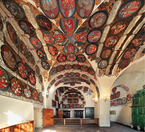 Interi r historick m stnosti s klenut m stropem zdoben m barevn mi erby a ornamenty, d ev n mi dve mi a zelen m kachlov m kamenem.  Popis generovan  AI