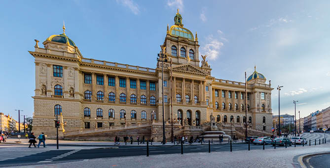 Velk historick  budova s bohatou architekturou a zelenou kopul  stoj  na n m st , kde chod  n kolik lid  a proj  d j  auta.  Popis generovan  AI