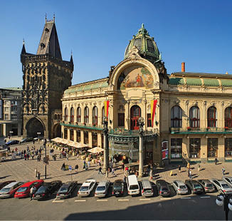 Historick budova s bohatou architekturou a v    stoj  na slune n m n m st , kde se pohybuje n kolik lid  a zaparkovan  jsou auta.  Popis generovan  AI