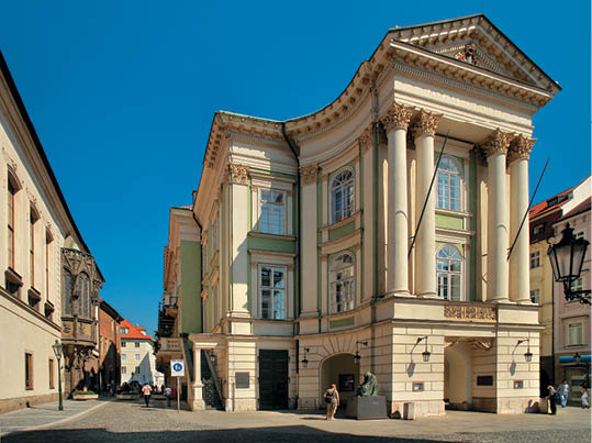 Historick budova s klasickou architekturou a sloupy stoj  na rohu dl  d n  ulice, kde se pohybuj  lid  v b  n m oble en  za slune n ho dne.  Popis generovan  AI