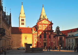 N m st s historickou architekturou v etn  dvou v    a  erven  budovy osv cen  sluncem, kde se pohybuje n kolik lid .  Popis generovan  AI