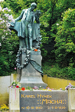 Socha osoby v historick m od vu dr c  kv tiny stoj c  na podstavci s n pisem Karel Hynek M cha obklopen  zelen  a kv tinami.  Popis generovan  AI