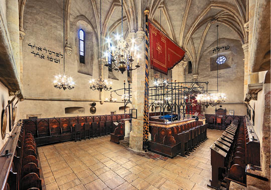 Interi r historick synagogy s d ev n mi lavicemi, kamennou podlahou, gotick mi klenbami a z v sn mi lustry.  Popis generovan  AI