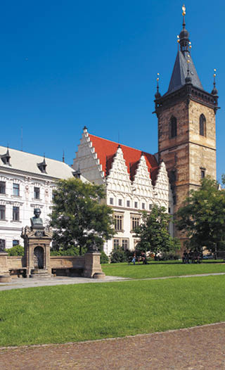 Historick budova s v    a sochou na n m st  s tr vn kem a stromy za slune n ho dne.  Popis generovan  AI