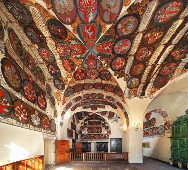 Interi r historick m stnosti s klenut m stropem zdoben m barevn mi erby a ornamenty, d ev n mi dve mi a zelen m kachlov m kamenem.  Popis generovan  AI