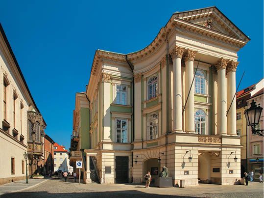 Historick budova s klasickou architekturou a sloupy stoj  na rohu dl  d n  ulice, kde se pohybuj  lid  v b  n m oble en  za slune n ho dne.  Popis generovan  AI
